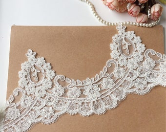 May include: White lace trim featuring a delicate floral design and beaded embellishments. The trim is displayed against a brown backdrop.