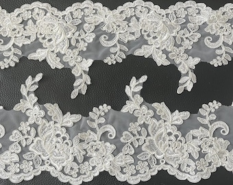 May include: Close-up of two strips of white floral lace. The lace features a delicate pattern of flowers, leaves, and intricate details. The lace is set against a dark background, highlighting the intricate design and texture.