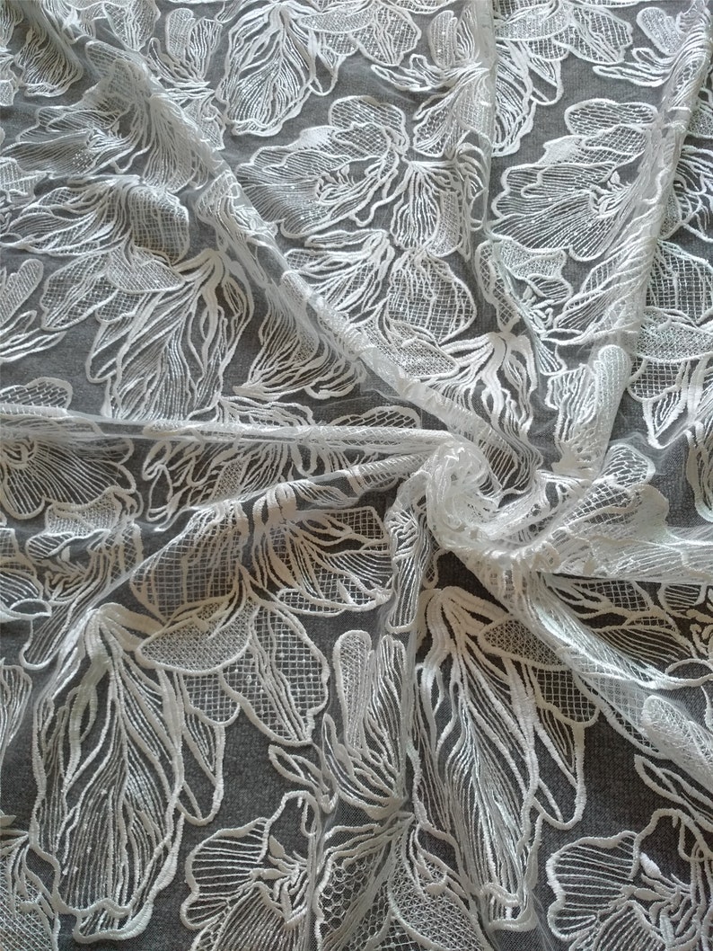 May include: White floral lace fabric with a delicate, intricate pattern. The fabric is sheer and has a soft, flowing texture.