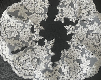 May include: Ivory floral lace trim with a scalloped edge. The delicate lace features detailed floral patterns and is likely used for embellishing garments or crafts. The lace is displayed against a dark background.