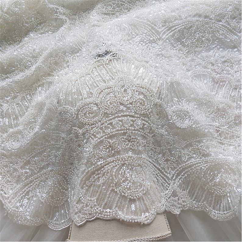 May include: White beaded lace fabric with a delicate floral pattern. The fabric is sheer and has a soft, flowing texture.