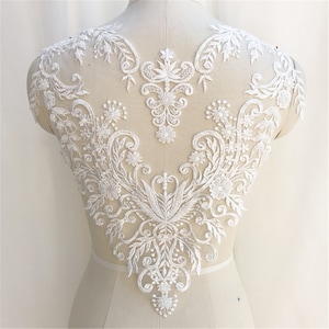 May include: White lace applique featuring floral and scroll designs. The lace is delicate and intricate, with a scalloped edge. This applique is ideal for adding a touch of elegance to any garment.
