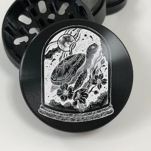 May include: Black metal grinder with a white design of a turtle inside a glass dome with a moon and stars.