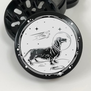 Puede incluir: Molino de caf&eacute; negro y blanco con un dachshund con un casco espacial flotando en el espacio. El molino tiene un dise&ntilde;o circular con fondo blanco y detalles negros.