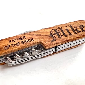 May include: A wooden multi-tool with a stainless steel blade and tools. The handle is engraved with the name "Mike" in an ornate font and the text "FATHER OF THE BRIDE".