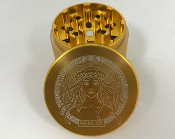 May include: Gold metal grinder with a Medusa design. The grinder has a circular top with a detailed engraving of Medusa's face and snakes. The text "MEDUSA" is engraved below the image.