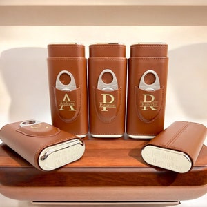 May include: Five brown leather cigar cases with silver accents and personalized engravings. Each case has a built-in cutter and a silver band with a name and date.