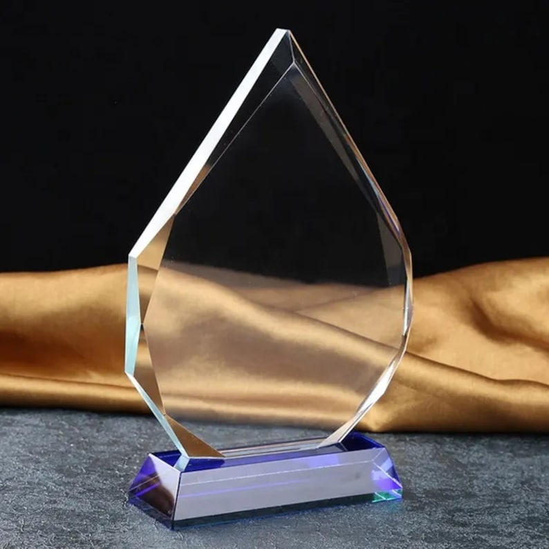 May include: A clear glass award with a blue base. The award is shaped like a teardrop and has a flat surface for engraving.