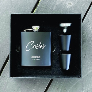 Pode incluir: Uma caixa de presente preta com uma garrafa de licor preta, dois copos de shot e um funil. A garrafa de licor é gravada com o nome "Carlos", "Groomsman" e a data "11.11.2021".