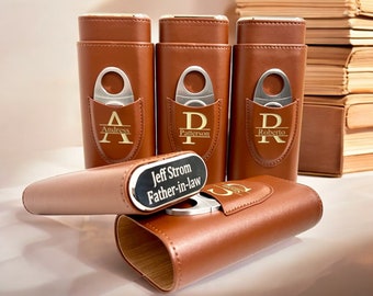 May include: Five brown leather cigar cases with gold lettering and a silver cigar cutter. The cases are personalized with names and initials. The cases are arranged in a row on a white surface.
