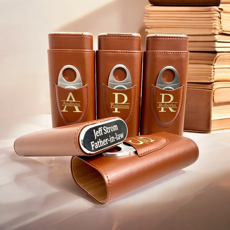 May include: Five brown leather cigar cases with gold lettering and a silver cigar cutter. The cases are personalized with names and initials. The cases are arranged in a row on a white surface.