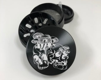 May include: Black metal grinder with a design of two skulls with mushrooms growing out of the top. The grinder has a black lid and a black base.
