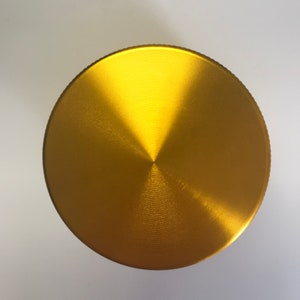 Puede incluir: Una tapa de molinillo de metal de color dorado, de forma circular y con un acabado cepillado.