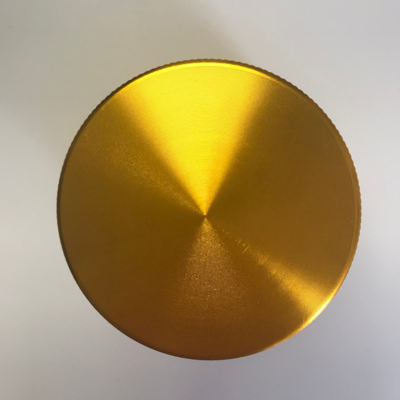 Puede incluir: Una tapa de molinillo de metal de color dorado, de forma circular y con un acabado cepillado.