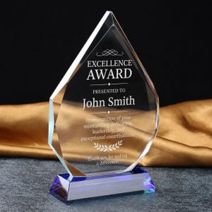 May include: A clear, diamond-shaped glass award trophy featuring a blue glass base. The text reads: EXCELLENCE AWARD, PRESENTED TO, John Smith, In recognition of your outstanding dedication, leadership, and exceptional contributions, Thank you for making a difference.