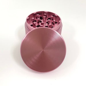 Puede incluir: Un molinillo de metal rosa con acabado cepillado. El molinillo tiene una tapa y un compartimento para moler hierbas.