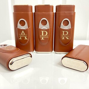 May include: Five brown leather cigar cases with silver accents. Each case has a personalized inscription and a built-in cigar cutter. The inscriptions include "Andress", "Patterson", "Roberto", "Austin + Risa 06.18.2022", and "Jeff Strom Father-in-law".
