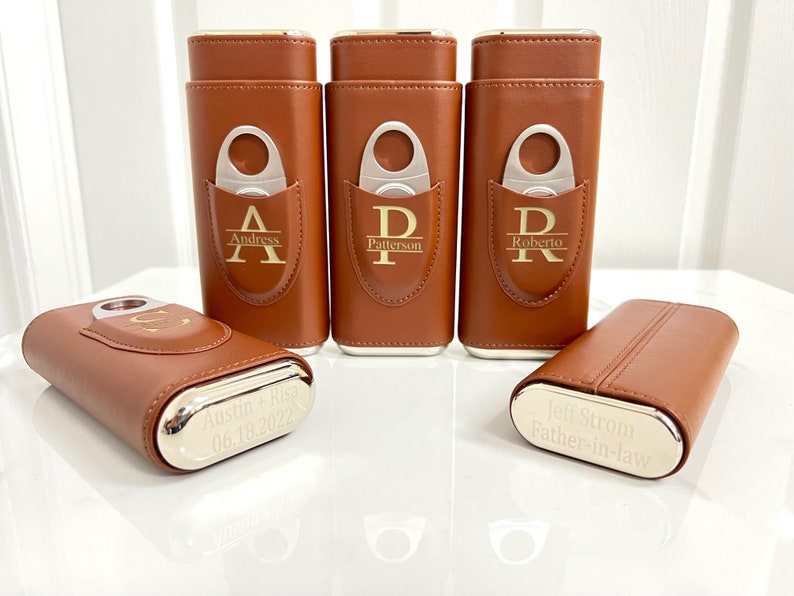 May include: Five brown leather cigar cases with silver accents. Each case has a personalized inscription and a built-in cigar cutter. The inscriptions include "Andress", "Patterson", "Roberto", "Austin + Risa 06.18.2022", and "Jeff Strom Father-in-law".