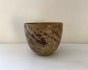 May include: A handcrafted ceramic bowl with a textured, earthy appearance. The bowl features a light brown base with dark brown splatters and diagonal lines. The interior is a darker shade of brown. The bowl sits on a white surface.
