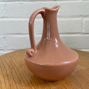 May include: A small, peach-colored ceramic pitcher with a curved handle and a spout. The pitcher has a rounded base and a smooth, glossy finish. It is displayed on a wooden surface.