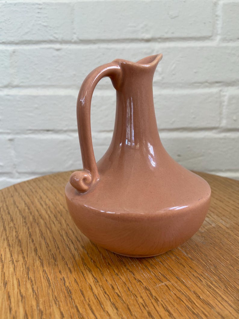 May include: A small, peach-colored ceramic pitcher with a curved handle and a spout. The pitcher has a rounded base and a smooth, glossy finish. It is displayed on a wooden surface.