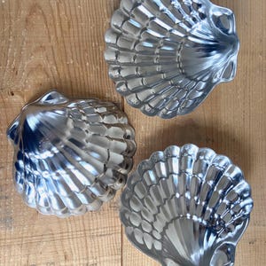 May include: Three shiny, silver-colored seashell-shaped molds. The molds are made of metal and have detailed ridges and curves, resembling real seashells. They are arranged on a light brown wooden surface.