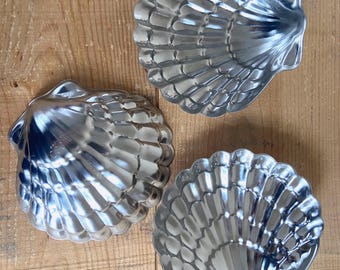 May include: Three shiny, silver-colored seashell-shaped molds. The molds are made of metal and have detailed ridges and curves, resembling real seashells. They are arranged on a light brown wooden surface.