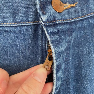 May include: Close-up of blue denim jeans with a copper-colored button and zipper. The zipper is partially open, revealing the metal teeth. The jeans have visible stitching and a Wrangler button.