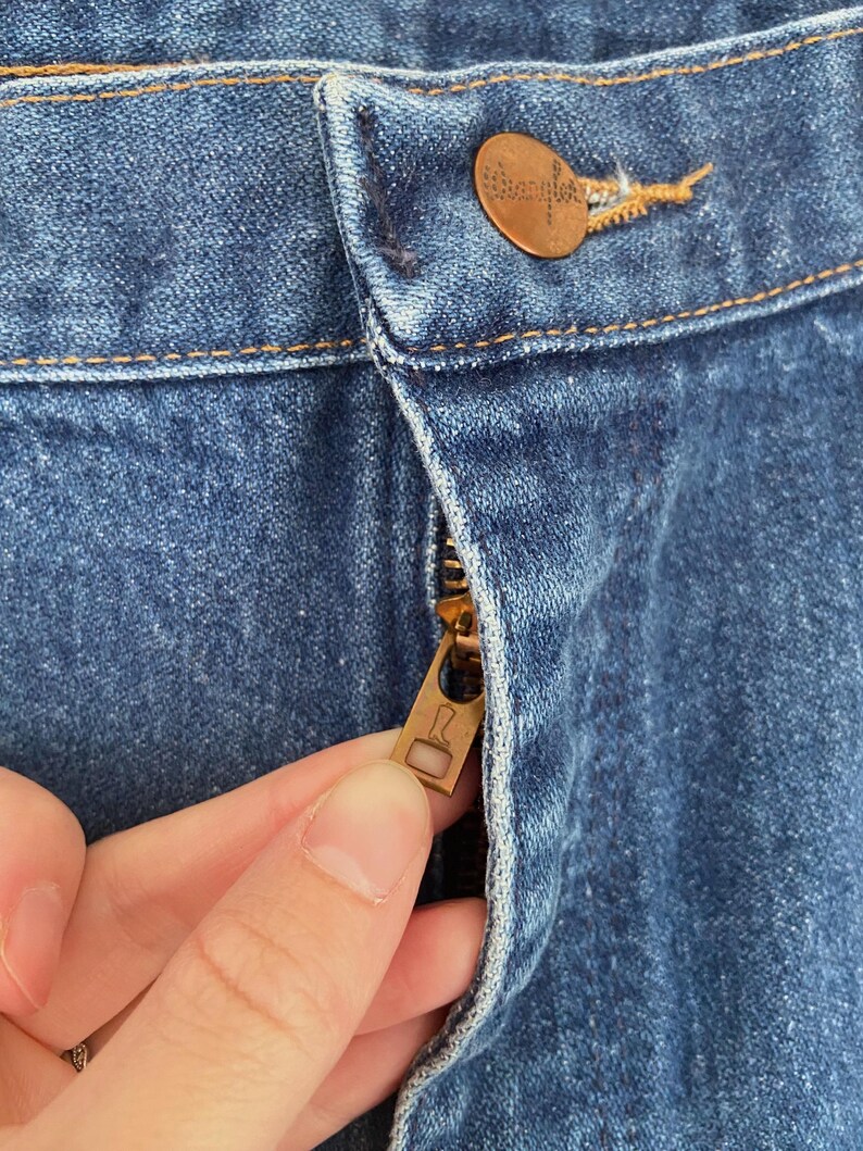 May include: Close-up of blue denim jeans with a copper-colored button and zipper. The zipper is partially open, revealing the metal teeth. The jeans have visible stitching and a Wrangler button.