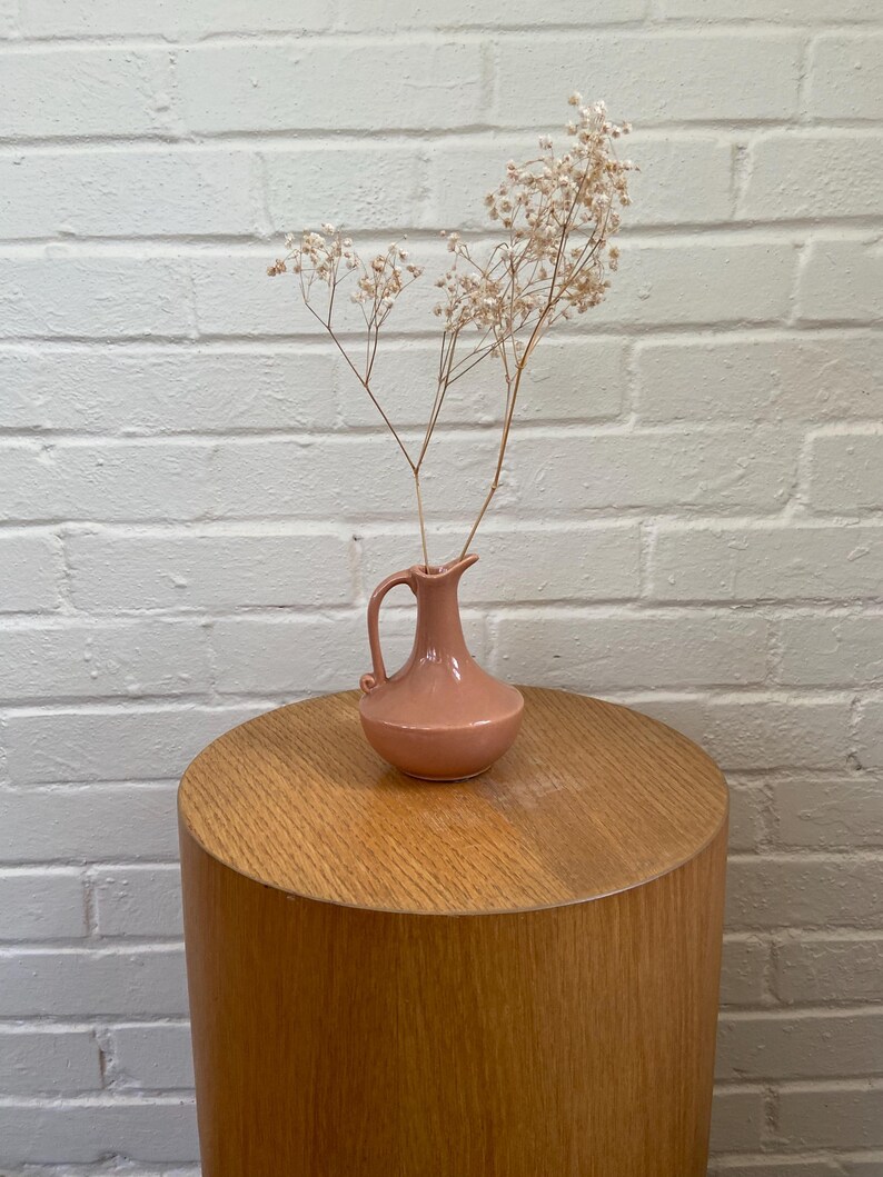 May include: A small, pink ceramic vase with a handle holds dried flowers. The vase sits on a round, wooden side table. The background is a white brick wall. The vase is a muted, dusty rose color.