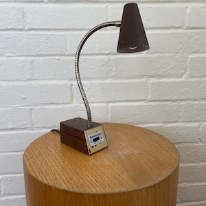 May include: A vintage desk lamp with a dark brown conical shade and a flexible, silver-toned gooseneck. The lamp is attached to a rectangular wooden base with the text "benson" and control buttons. It sits on a cylindrical wooden table.