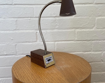 May include: A vintage desk lamp with a dark brown conical shade and a flexible, silver-toned gooseneck. The lamp is attached to a rectangular wooden base with the text "benson" and control buttons. It sits on a cylindrical wooden table.