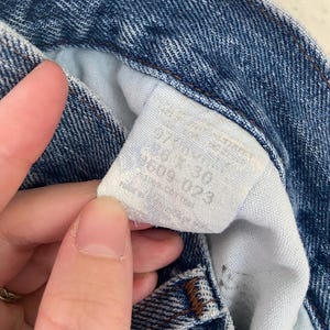 May include: Close-up of blue denim jeans with a white tag. The tag displays the size as 28 x 30. The jeans are made of 100% cotton. The denim has a classic, worn look.