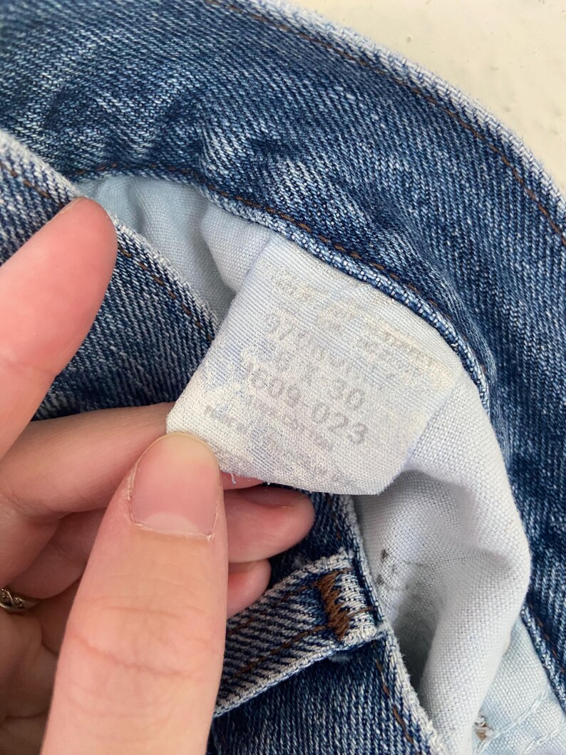 May include: Close-up of blue denim jeans with a white tag. The tag displays the size as 28 x 30. The jeans are made of 100% cotton. The denim has a classic, worn look.