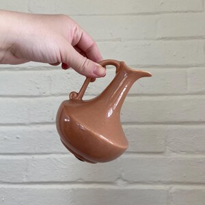 May include: A peach-colored ceramic pitcher with a curved handle and a spout. The pitcher has a bulbous base and a smooth, glossy finish. The handle has a small decorative swirl. The pitcher is held against a white brick wall.