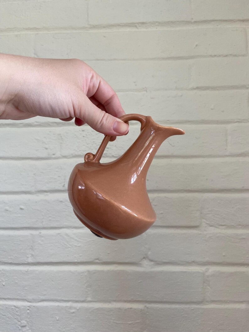 May include: A peach-colored ceramic pitcher with a curved handle and a spout. The pitcher has a bulbous base and a smooth, glossy finish. The handle has a small decorative swirl. The pitcher is held against a white brick wall.