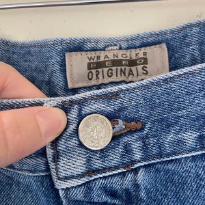 May include: Close-up of blue denim jeans with a button closure. The jeans feature a "Wrangler Hero Originals" label. The button is silver with the Wrangler logo. The denim has a classic, worn look.
