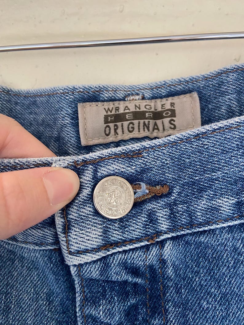 May include: Close-up of blue denim jeans with a button closure. The jeans feature a "Wrangler Hero Originals" label. The button is silver with the Wrangler logo. The denim has a classic, worn look.