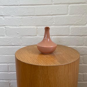 May include: A small, peach-colored ceramic vase with a rounded base and a slender neck. The vase sits atop a cylindrical wooden side table. The background is a white brick wall. The vase is a decorative item.