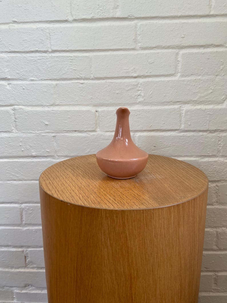 May include: A small, peach-colored ceramic vase with a rounded base and a slender neck. The vase sits atop a cylindrical wooden side table. The background is a white brick wall. The vase is a decorative item.