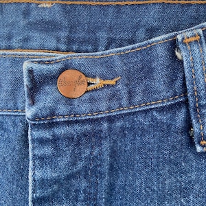 May include: Close-up of blue denim jeans with a copper-colored button. The button is embossed with the brand name "Wrangler". The jeans have a classic five-pocket design and are stitched with a contrasting yellow thread.