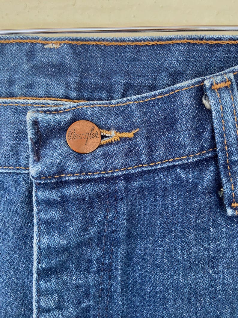 May include: Close-up of blue denim jeans with a copper-colored button. The button is embossed with the brand name "Wrangler". The jeans have a classic five-pocket design and are stitched with a contrasting yellow thread.