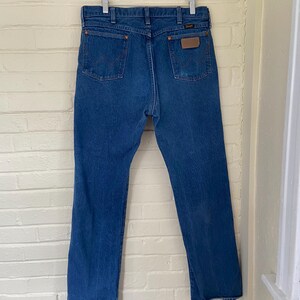 May include: Blue denim jeans hanging on a wooden hanger. The jeans have a classic five-pocket design with a brown leather patch on the back. The Wrangler logo is visible on the patch. The jeans are a medium wash and appear to be vintage.