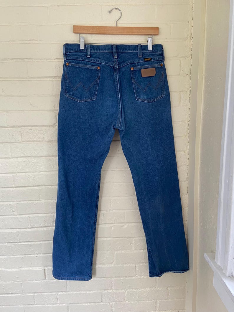 May include: Blue denim jeans hanging on a wooden hanger. The jeans have a classic five-pocket design with a brown leather patch on the back. The Wrangler logo is visible on the patch. The jeans are a medium wash and appear to be vintage.