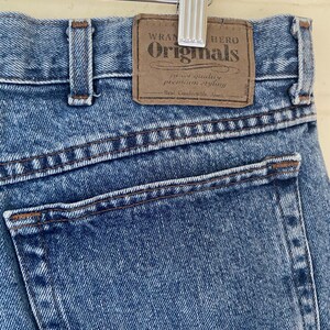 May include: Close-up of blue denim jeans with a brown leather patch. The patch reads "Wrangler Originals" with text below. The jeans are hanging on a silver hanger. The jeans have a classic five-pocket design.