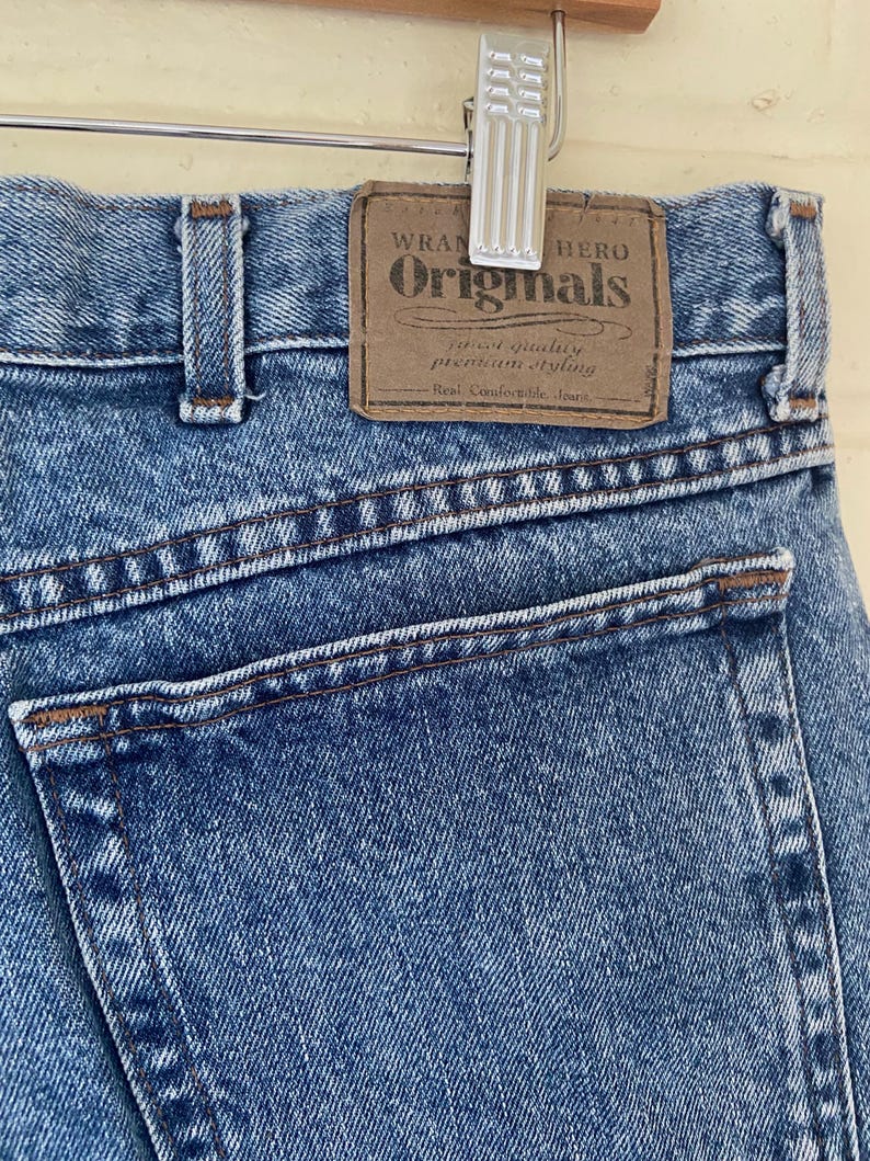 May include: Close-up of blue denim jeans with a brown leather patch. The patch reads "Wrangler Originals" with text below. The jeans are hanging on a silver hanger. The jeans have a classic five-pocket design.