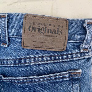 May include: Close-up of blue denim jeans with a brown leather patch. The patch reads "Wrangler Hero Originals" with additional text. The jeans have a back pocket and visible stitching.