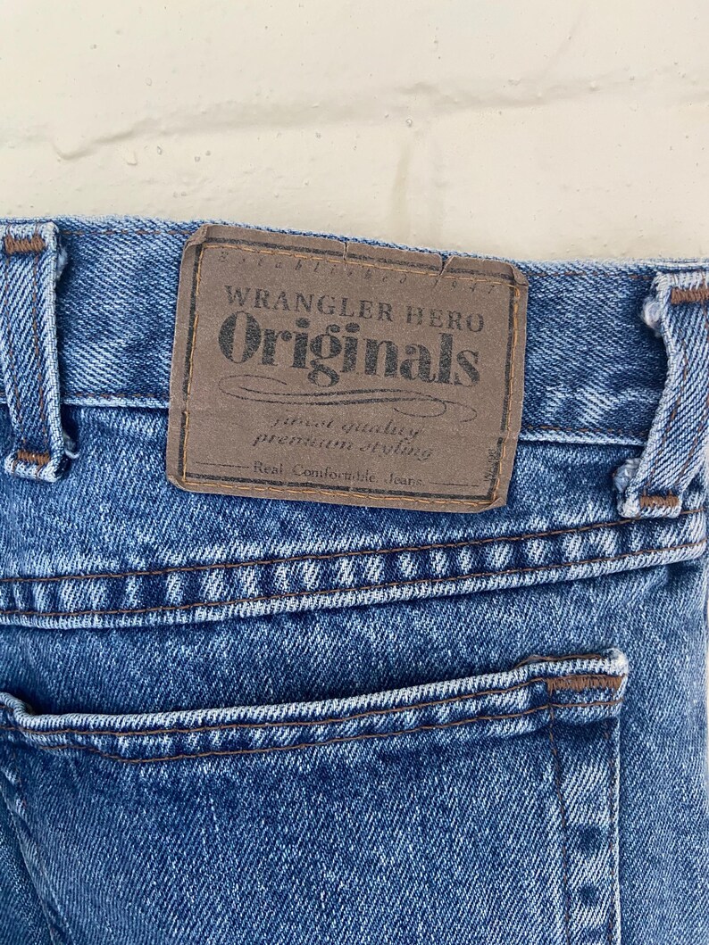 May include: Close-up of blue denim jeans with a brown leather patch. The patch reads "Wrangler Hero Originals" with additional text. The jeans have a back pocket and visible stitching.