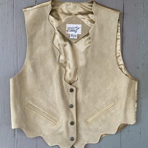 May include: A tan suede vest featuring a scalloped front edge and neckline, four metal buttons, and two welt pockets. The interior is lined with a matching tan satin fabric. A label inside reads Boot Country with a size L tag below it.