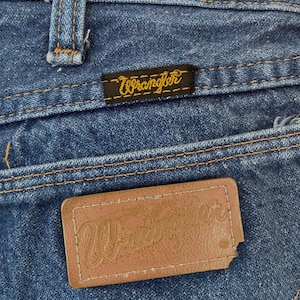 May include: Close-up of blue denim jeans with the brand name "Wrangler" visible on two separate tags. One tag is a small black rectangle with yellow stitching, and the other is a brown leather patch.